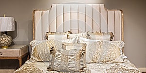 Elegant neutral satin bed cover and pillows against padded bed stead and beige wall with matching lamp