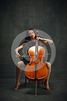 Elegant, passionate musician, beautiful woman in black dress sitting and playing cello against dark green vintage