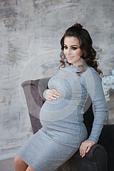 Elegant pregnant model in beautiful evening dress sit by the armchair. Background of bedroom