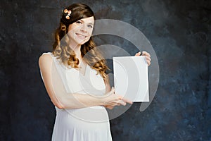 Elegant pregnant young woman wearing white dress holding white canvas in her hands, black wall on background.