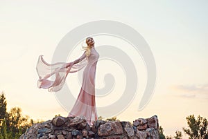 Elegant princess with blond fair wavy hair with tiara on it, wearing a long light pink rose fluttering dress, standing