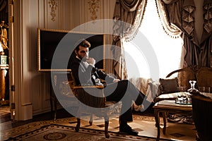 Elegant prosperous businessman in formal black suit, sits on chair in royal room, feels relaxed, has serious thoughtful