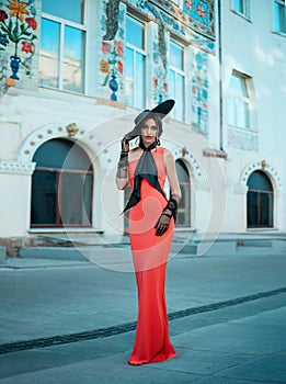 Elegant retro lady in a long red evening glamour dress. Young stylish trendy woman in a black hat with wide brim and