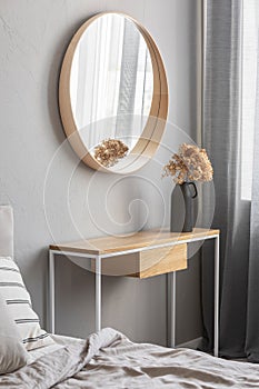 Elegant round mirror in wooden frame above fancy console table with flowers in vase in trendy bedroom interior with beige vase