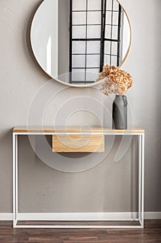 Elegant round mirror in wooden frame above fancy console table with flowers in vase in trendy bedroom interior with beige vase