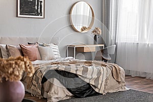 Elegant round mirror in wooden frame above fancy console table with flowers in vase in trendy bedroom interior with beige vase