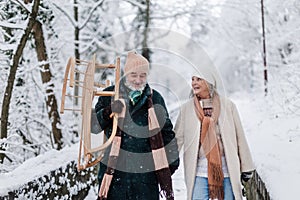 Elegant senior couple walking with sledge in the snowy park, during cold winter day. Winter vacation in the mountains