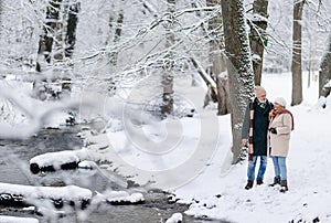 Elegant senior couple walking in the snowy park near the river, during winter snowy day. Winter vacation in the