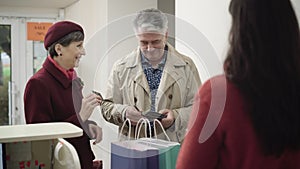 Elegant senior husband giving credit card to beautiful smiling wife in shop. Portrait of cheerful Caucasian couple