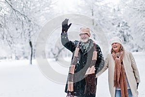 Elegant senior man waving to someone during walk in the snowy park. Winter vacation in the mountains. Wintry landscape.