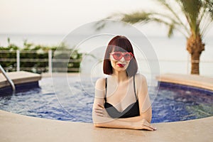 Elegant sexy woman with red hair in sunglasses and black bikini is posing near the swimming pool.