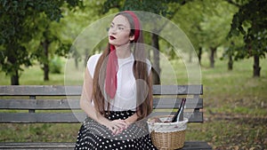 Elegant slim beautiful woman sitting on bench in summer park with picnic basket waiting for boyfriend. Portrait of