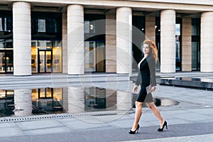 Elegant slim woman in black suit and high-heeled shoes, holding tablet in hands going for work. Businesswoman in motion. Confident