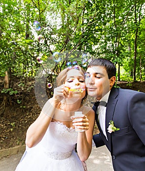 Elegant stylish groom and happy gorgeous bride have fun with bubble blower outdoors in park