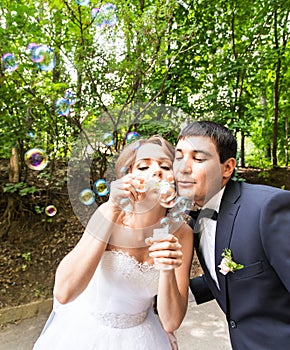 Elegant stylish groom and happy gorgeous bride have fun with bubble blower outdoors in park