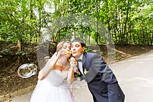 Elegant stylish groom and happy gorgeous bride have fun with bubble blower outdoors in park