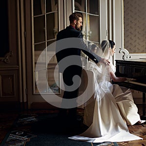 elegant stylish handsome groom hugging gently gorgeous bride while playing the fortepiano. unusual luxury wedding couple in retro photo