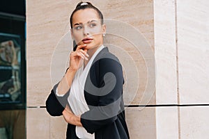 Elegant stylish lady stands in a black austere suit leaning on the wall of the business center