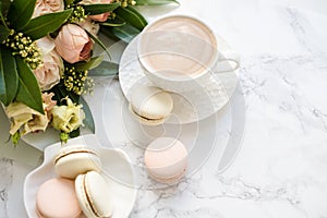 Elegant sweet dessert macarons, cup of coffee and pastel colored beige flowers bouquet on white marble