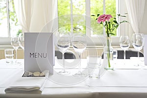 Elegant table setting, place with menu card, glasses, cutlery, napkin and a pink flower on a white tablecloth for a festive dinner