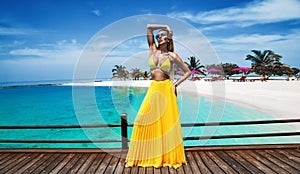 Elegant tanned woman in yellow bikini and long skirt on tropical beach on Maldives island. Summer fashion model is walking on the