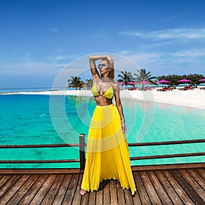 Elegant tanned woman in yellow bikini and long skirt on tropical beach on Maldives island. Summer fashion model is walking on the