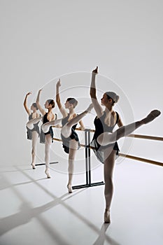 Elegant teen girls, ballerinas practicing, performing stretch at wooden barre against grey studio background.