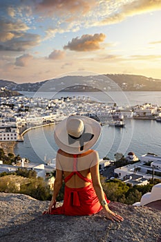 A elegant tourist woman in a red dress enjoys the view over the city of Mykonos island