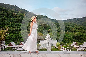 Elegant travel woman in hat and white dress walking in luxury resort with thai temple on background. Female traveler on