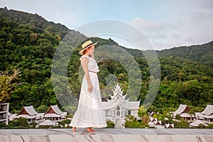Elegant travel woman in hat and white dress walking in luxury resort with thai temple on background. Female traveler on