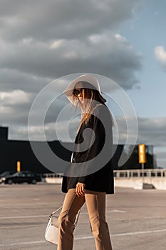 Elegant trendy young woman with bag in stylish casual clothes and straw hat walks to mall. Beautiful modern girl posing on parking