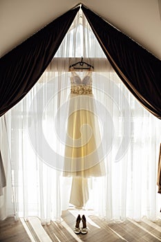 An elegant wedding dress hangs by a curtained window. Nearby are women's shoes