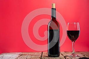 Elegant wine bottle and glass with red wine stands on the rustic wooden table on pink background. Space for text. Advertisement