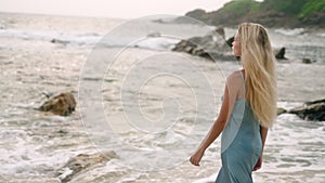 Elegant woman in blue dress strolls along sandy beach. Blonde hair, ocean waves, luxury solo travel. Freedom, relaxation