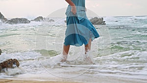 Elegant woman in blue sun dress strolls barefoot by sea. Serene female enjoys peaceful beach walk at sunset. Relaxing