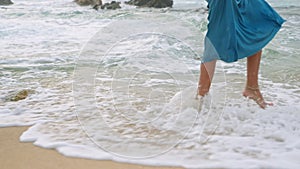 Elegant woman in blue sun dress strolls barefoot by sea. Serene female enjoys peaceful beach walk at sunset. Relaxing