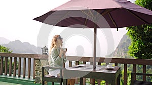 Elegant woman enjoys coffee on terrace with breathtaking mountain landscape. Morning refreshment in serene nature