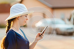 An elegant woman in a hat uses a smartphone. In the background, the house and car are blurred. Side view. The concept of social