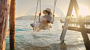 Elegant woman in long dress enjoying swinging on swing and looking on the sunset at sea beach. Tourism, summertime