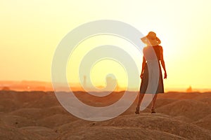 Elegant woman in long dress with straw hat standing on sand in desert, back view. Sunrise, backlight photo