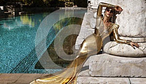 Elegant woman in long golden evening dress is posing outdoor in luxury resort in Turkey. Female model in amazing long dress. Vogue