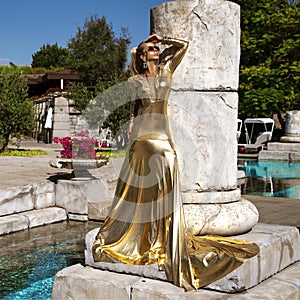 Elegant woman in long golden evening dress is posing outdoor in luxury resort in Turkey. Female model in amazing long dress. Vogue