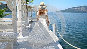 Elegant woman in long white dress standing on wooden pier and looking at the calm sea waves