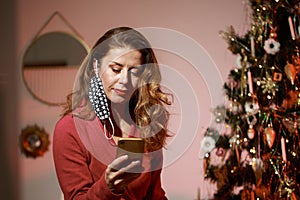 Elegant woman in a red cocktail dress, wearing protective face mask by the christmas tree, alone, lonely, talking and writing on