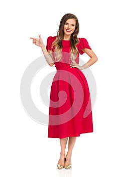 Elegant Woman In Red Dress And Gold High Heels Is Looking At Camera, Smiling And Pointing