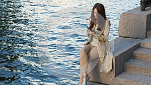 Elegant woman with smart phone sitting near river quay