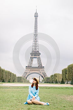 Elegant woman in straw hat and blue dress sitting on green grass in a park with Eiffel Tower on the background