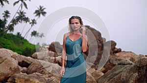 Elegant woman in teal dress stands by sea, rocky terrain, palm backdrop. Serene ambiance in natural pool setting