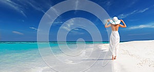 Elegant woman in white dress holds sun hat and walks on idyllic beach of maldives