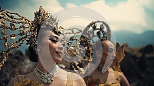 Elegant women in ornate costumes with golden headpieces at a cultural festival with a mountainous backdrop